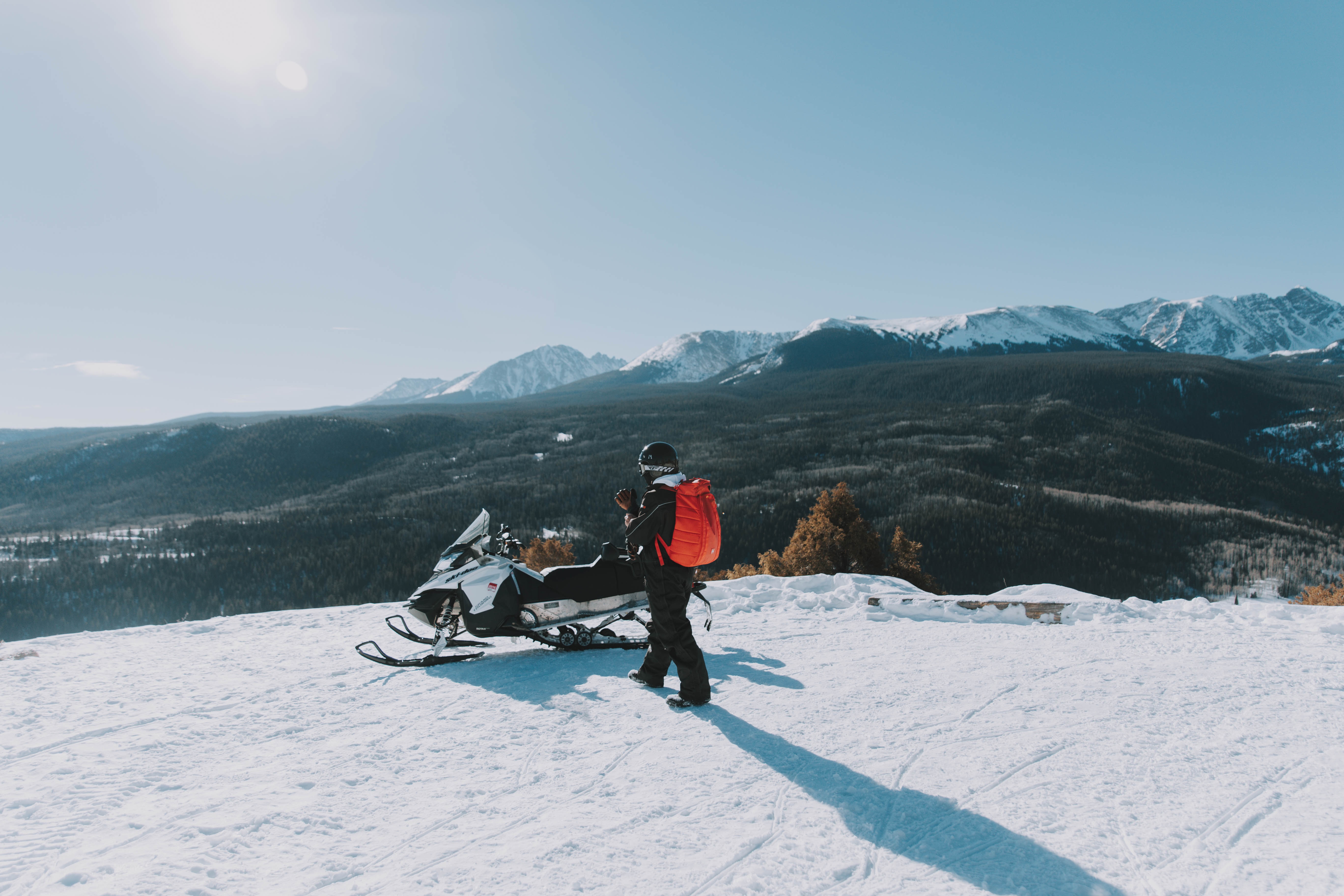 Man walks toward a snowmobile parked on a ridge overlooking a vast wilderness with mountains in the background